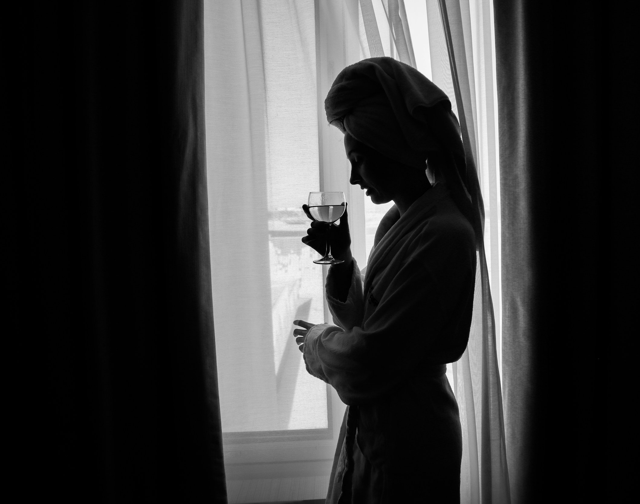 Silhouette of a woman with a towel wrapped around her head, holding a glass of water or wine, standing by a window with curtains.