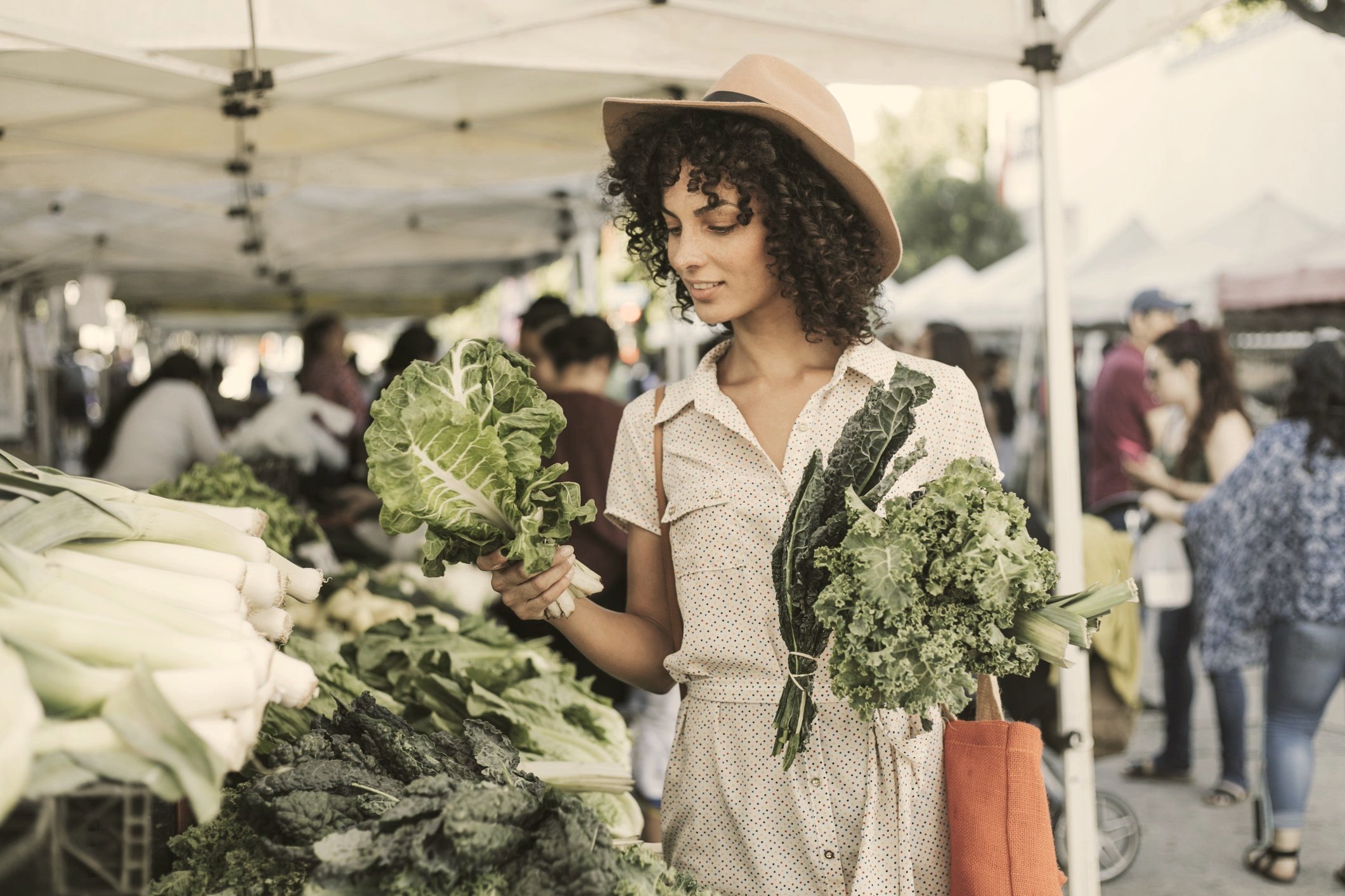 A woman shopping for vegetables at an outdoor market, holding leafy greens in each hand and examining a lettuce.