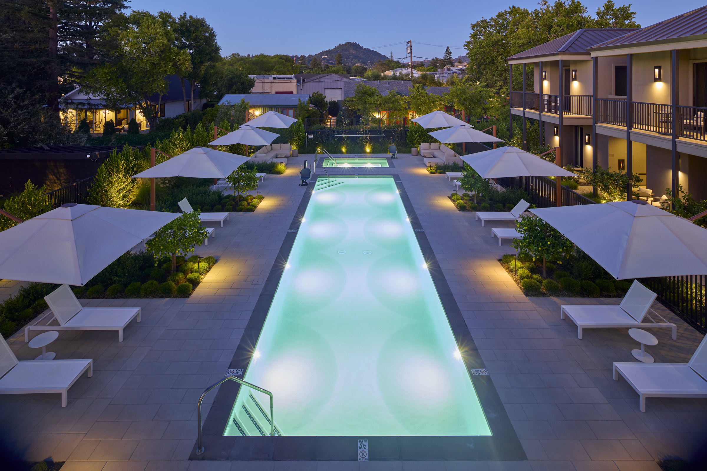 The pool courtyard at dusk, framed by white umbrellas, chaise lounges and the guest building balconies.