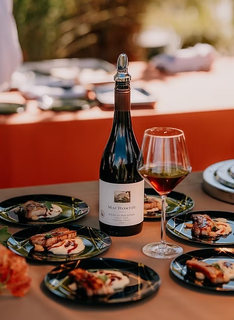 An outdoor table set with black plates of seared fish, a bottle of MacRostie Pinot Noir and a filled wine glass.