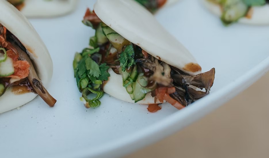 Steamed bao buns filled with roasted mushrooms, cucumber, tomato and herbs on a pale ceramic platter.